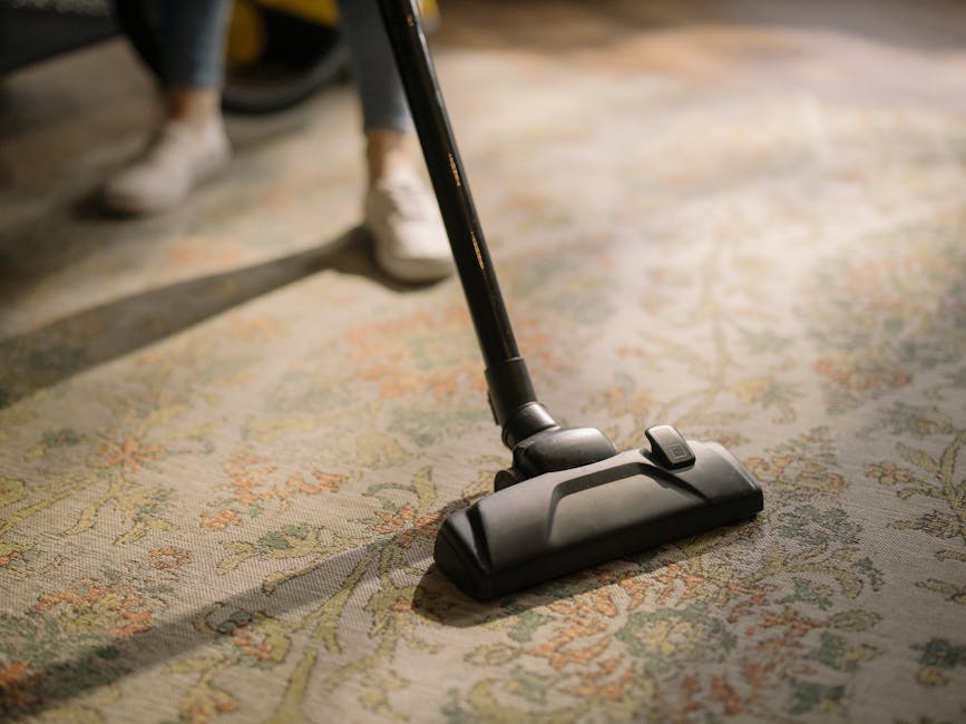 A woman with curly hair, wearing a beige jacket, dark jeans, and white shoes, is using a yellow carpet cleaning machine with a black hose and hand attachment to clean a patterned area rug in a living 