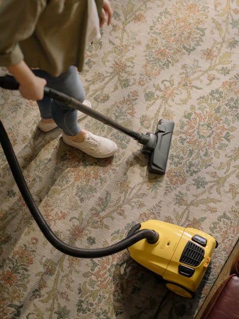 A person using a vacuum cleaner with a yellow canister and black hose is performing surface cleaning on an ornate, patterned area rug with floral motifs in a room. The individual, dressed in casual cl